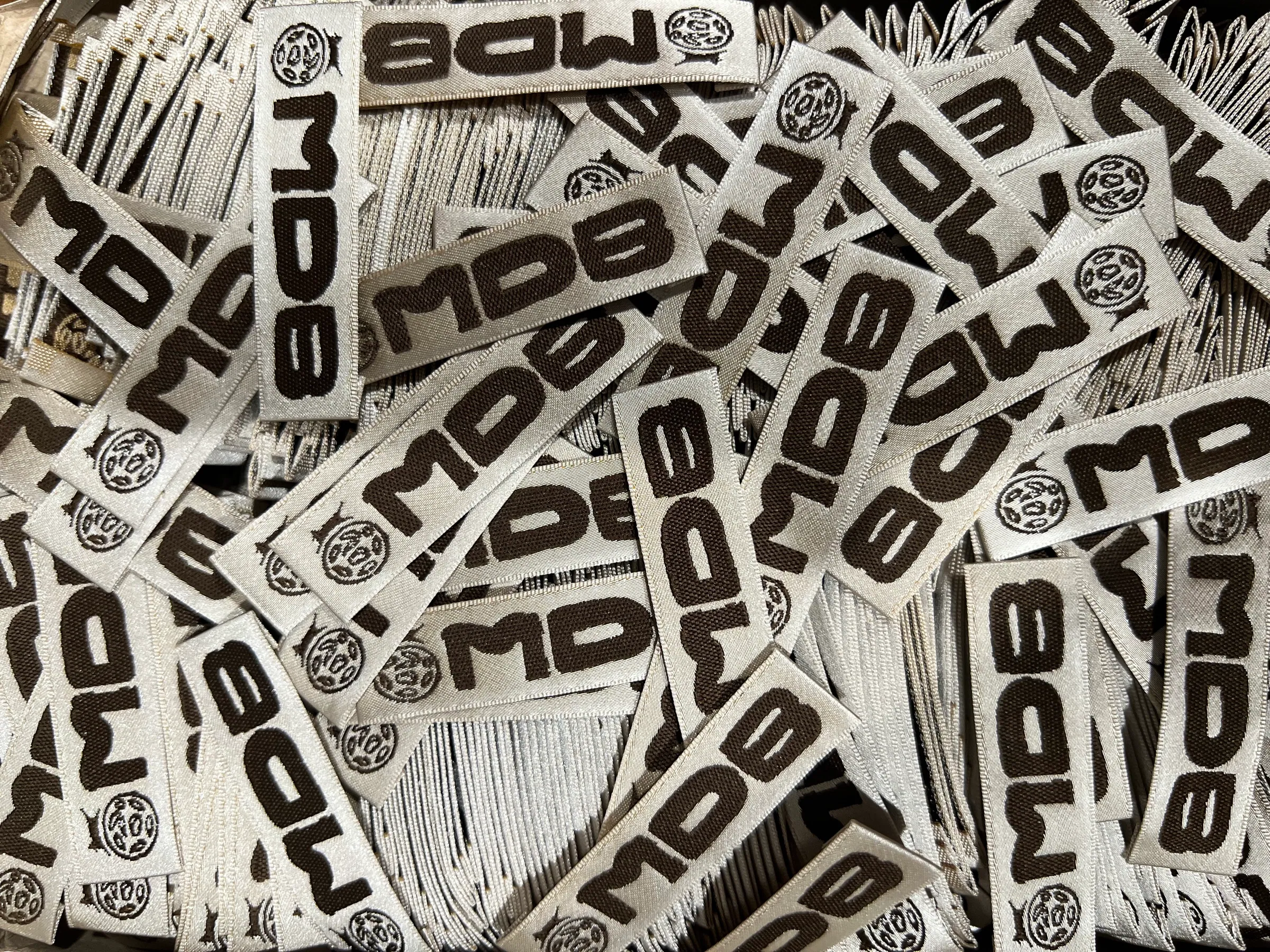 Woven labels by the hundred, spilled across a table. MDB and the cookie roundel woven in brown thread on a cream satin ground.