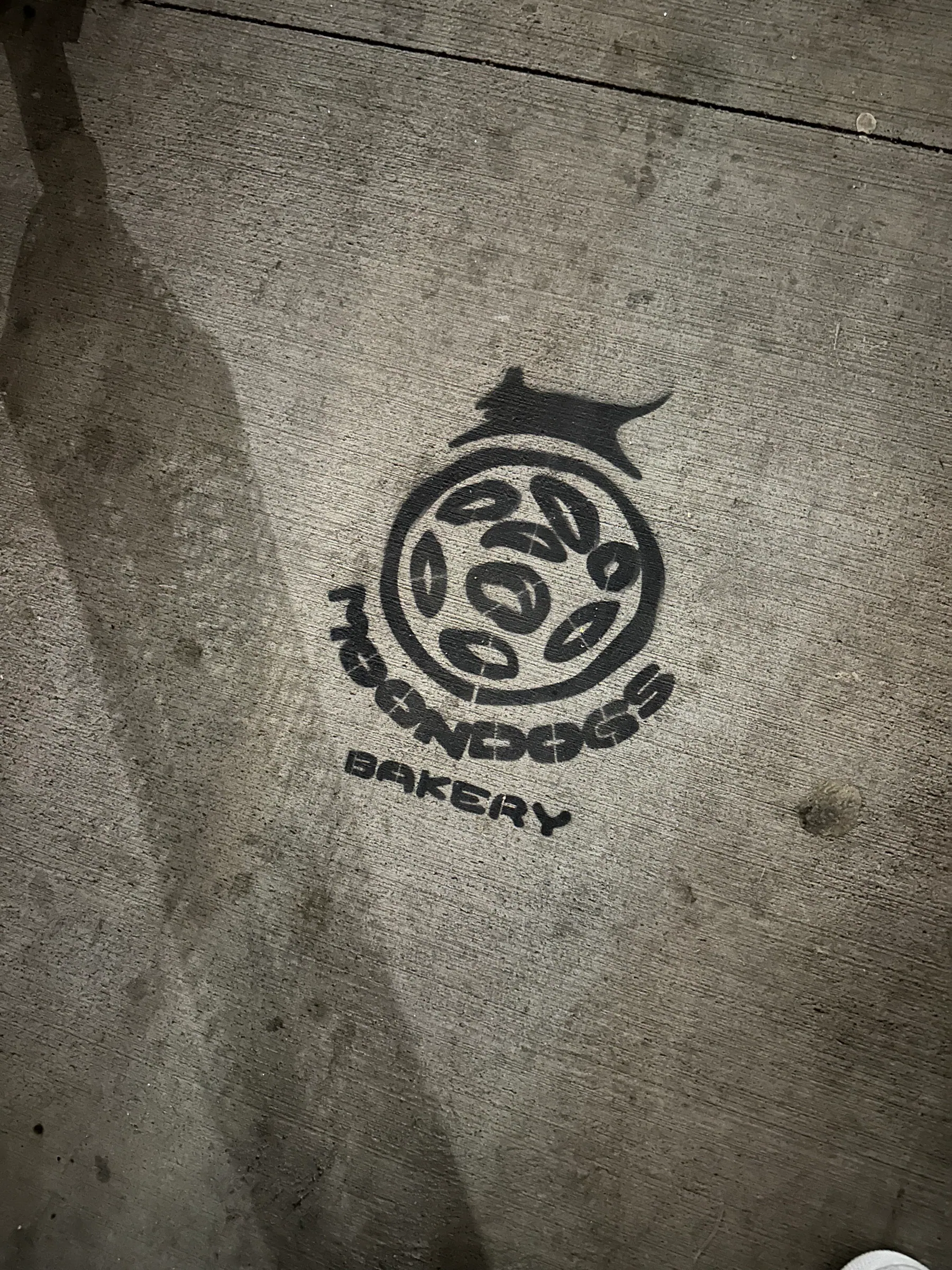 The mark stencilled in black spray paint on a concrete sidewalk, the dog leaping over the cookie roundel with MOONDOGS BAKERY curved around it.